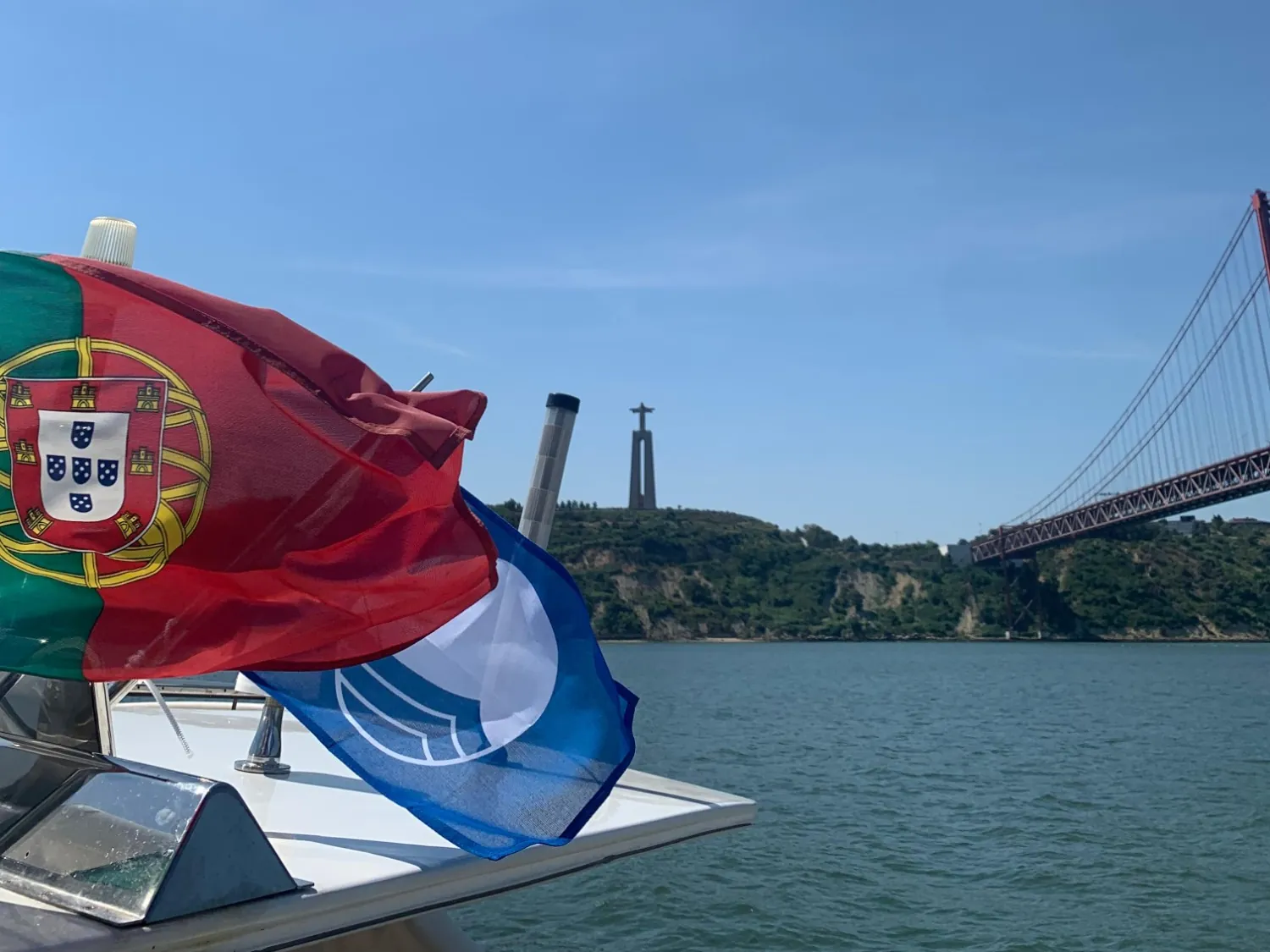 The Portuguese flag and the Blue Flag flying at the bow, with the Cristo Rei statue and the 25 de Abril Bridge across the Tagus